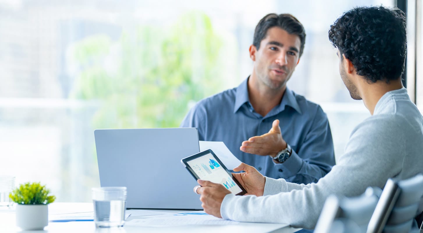 Two men engaged in conversation while sitting at a table in front of their laptop and tablet.
